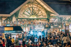Crowded Singapore hawker market at night, with numerous people walking among food stalls and bright lights.