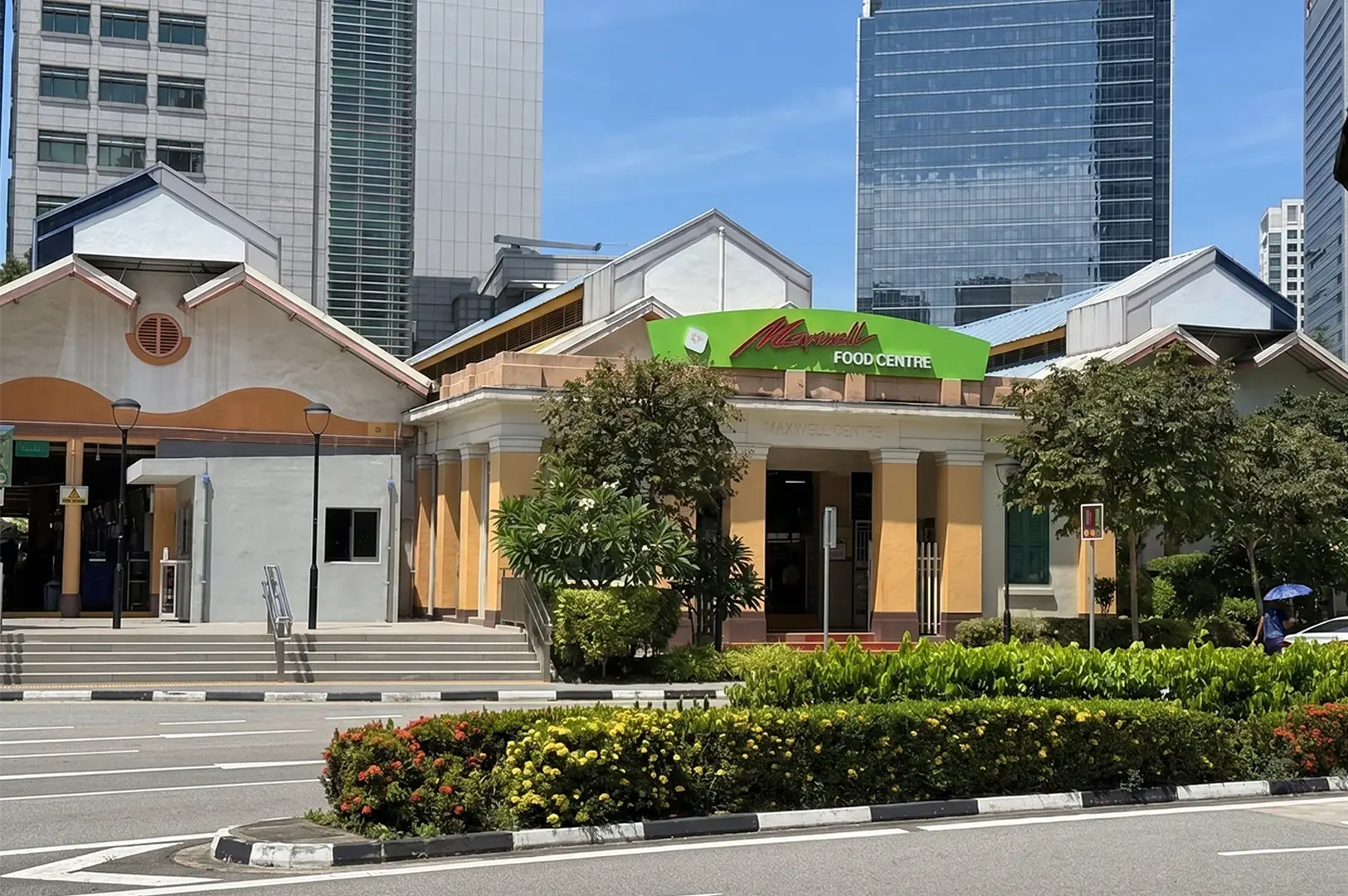Exterior view of Maxwell Food Centre, a famous hawker centre in Singapore, showing its distinctive yellow and white architecture against a backdrop of modern skyscrapers.