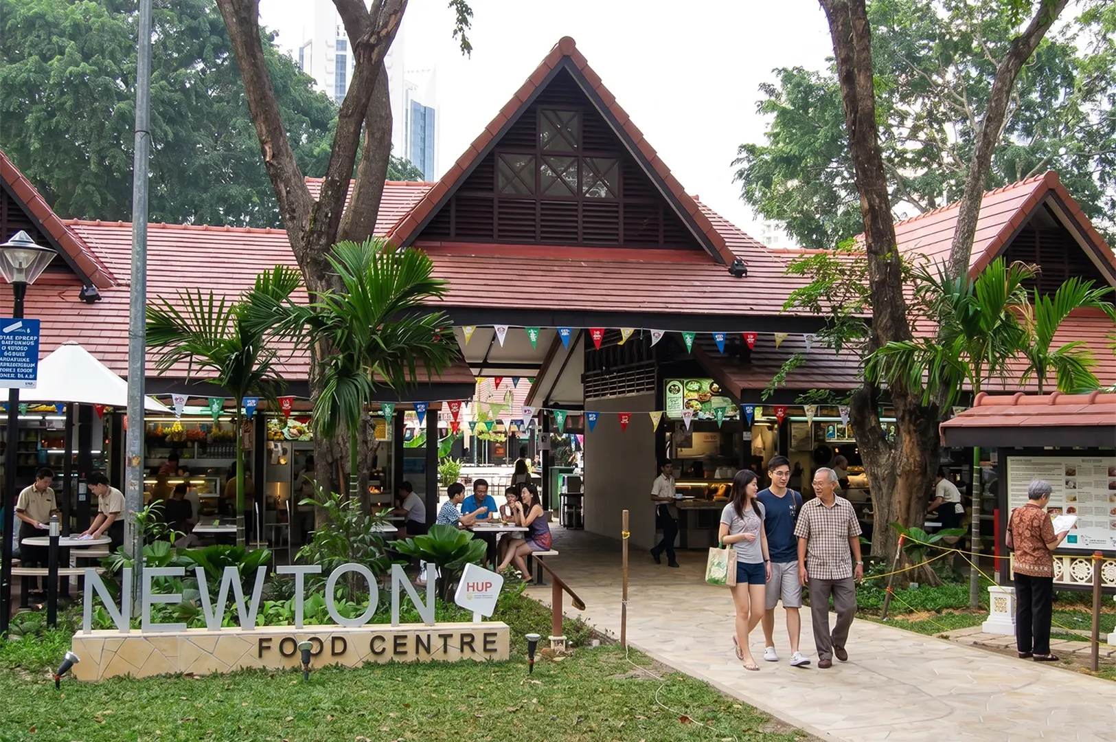 Entrance to Newton Food Centre in Singapore, featuring its iconic colonial-style orange roof, lush greenery, and people walking near the outdoor seating area.