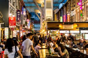 A bustling Singapore hawker street filled with people enjoying food and drinks at various stalls.