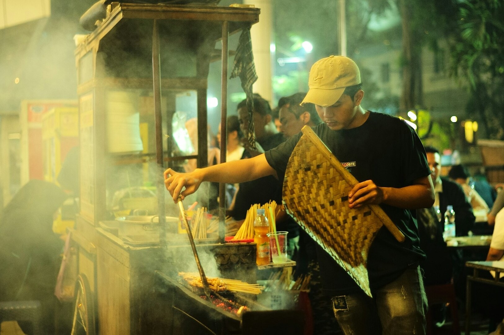A man cooks on a grill at night in a lively Singapore Hawker Centre, with smoke rising and food aromas filling the air.
