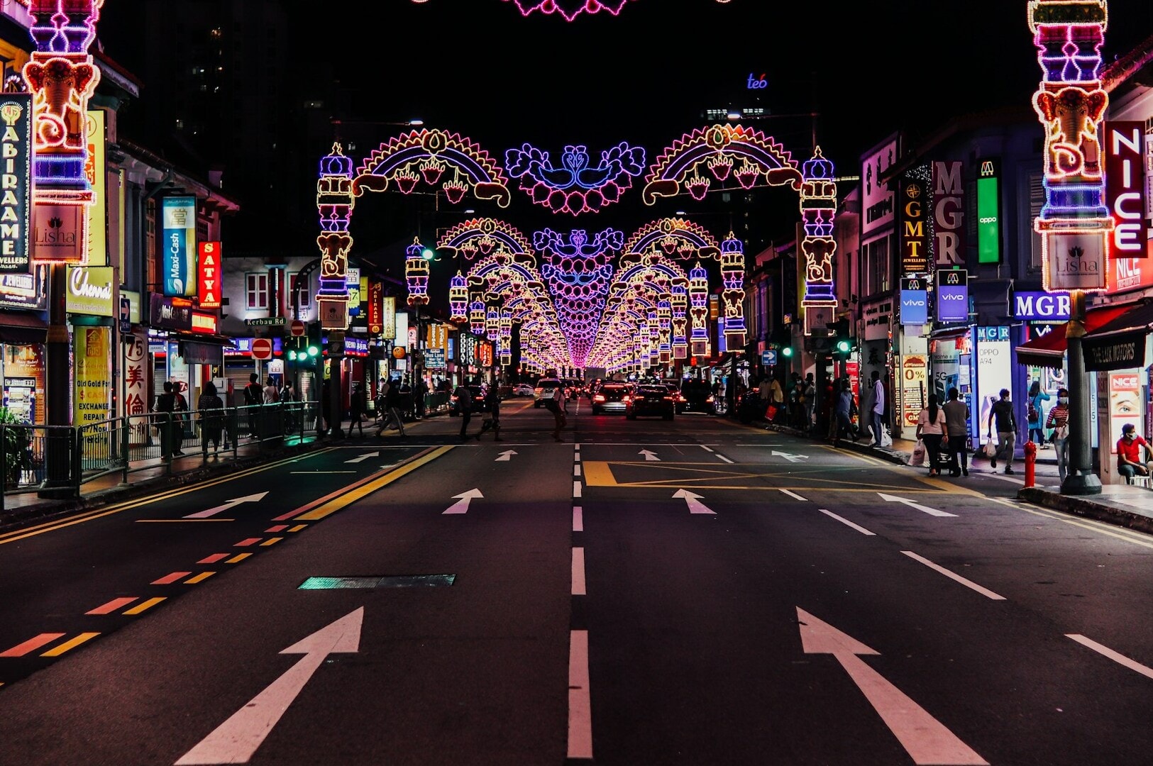 A lively street scene in Little India, Singapore, showcasing bright lights and numerous cars in motion.