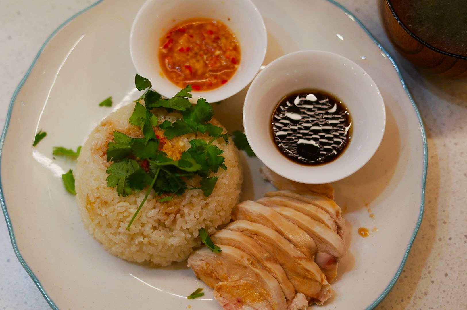 A plate of Hainanese chicken rice with sliced chicken, garnished rice, two dipping sauces in small bowls, offering a savory, appetizing appearance.