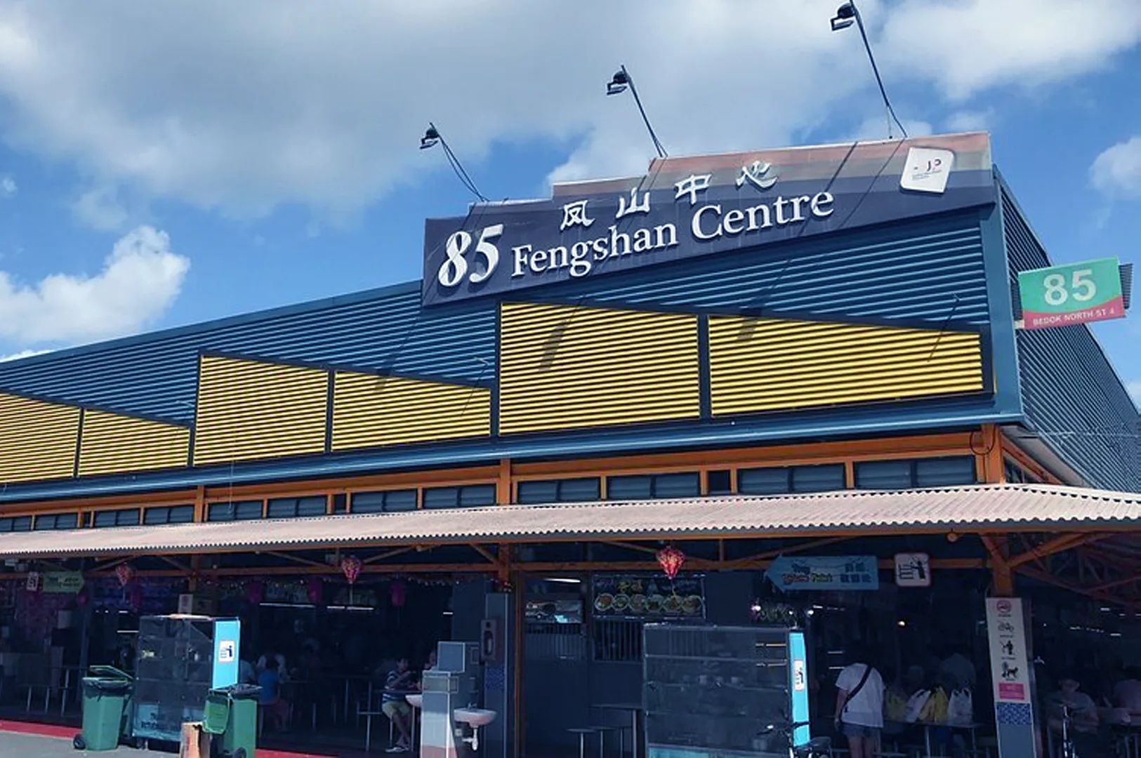 Facade of 85 Fengshan Centre under a blue sky, featuring a large sign with English and Chinese text, yellow paneling, and open-air dining area."