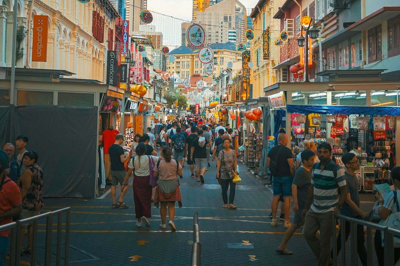 A bustling street scene in a vibrant market with people walking between stalls. Lanterns and decorations hang above, creating a lively, festive atmosphere.