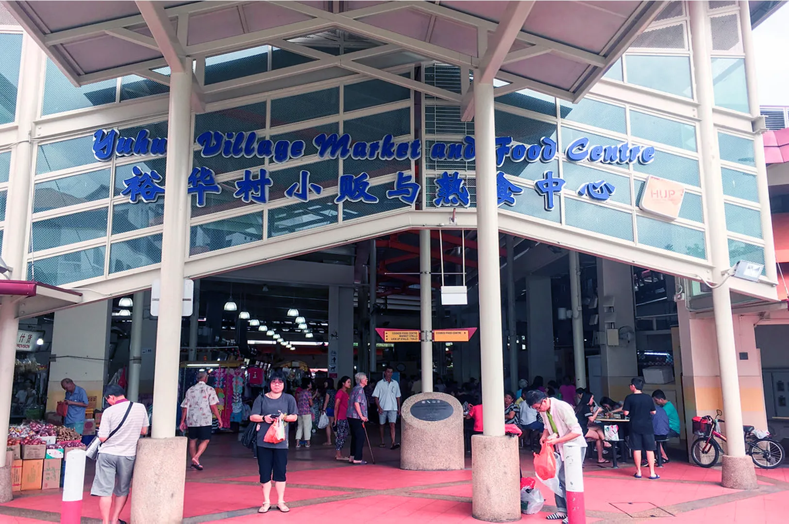Entrance of Yuhua Village Market and Food Centre, with people walking and gathered, vibrant atmosphere. Signage includes English and Chinese text.