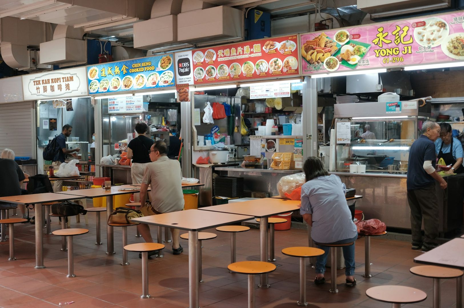 Indoor scene of a bustling food court with several open food stalls. Customers are seated at shared tables while others order food. The atmosphere is lively.