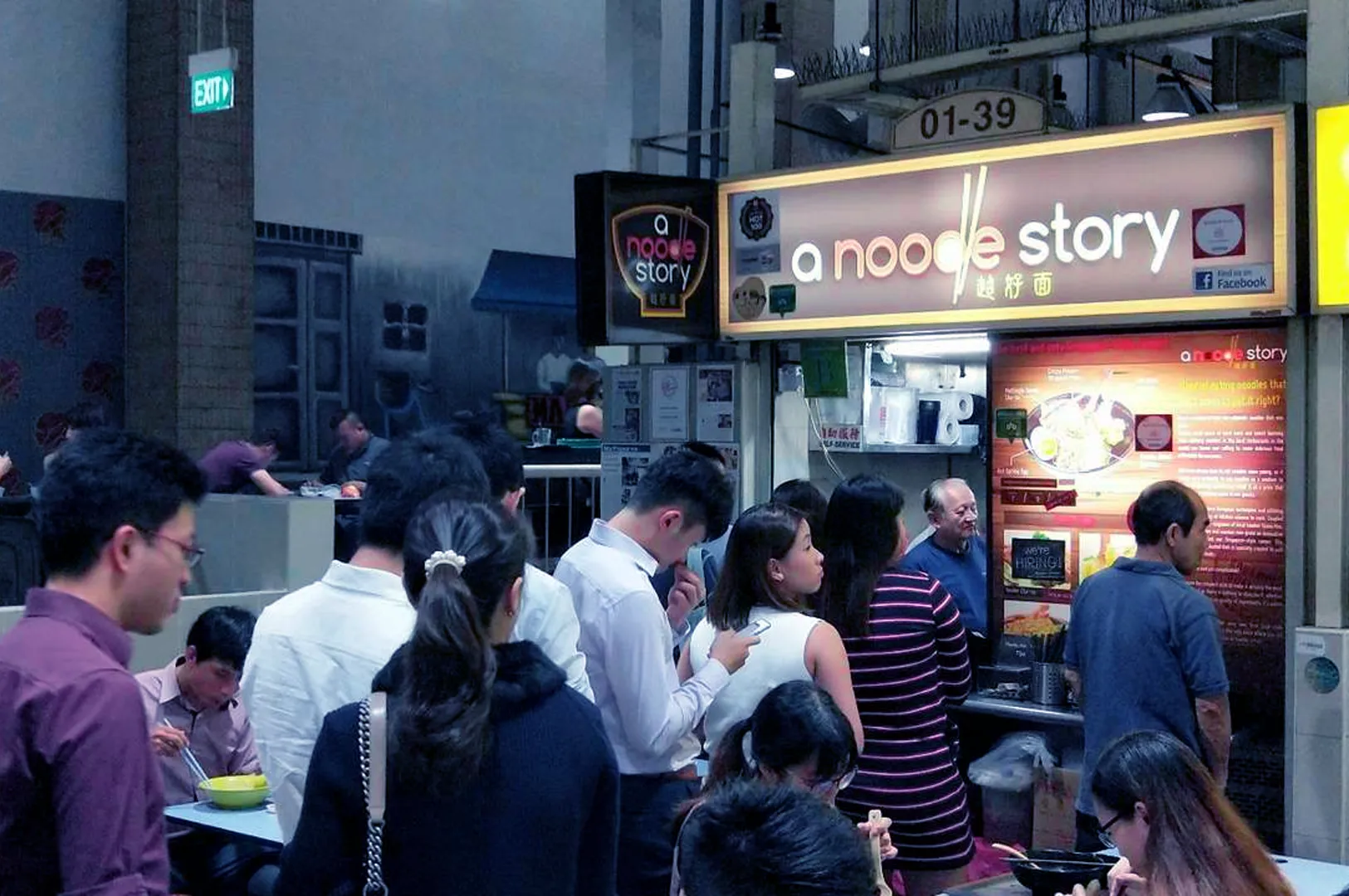 Queue of people at "A Noodle Story" food stall, a bustling hawker center. Customers eagerly await orders, creating an inviting, busy atmosphere.