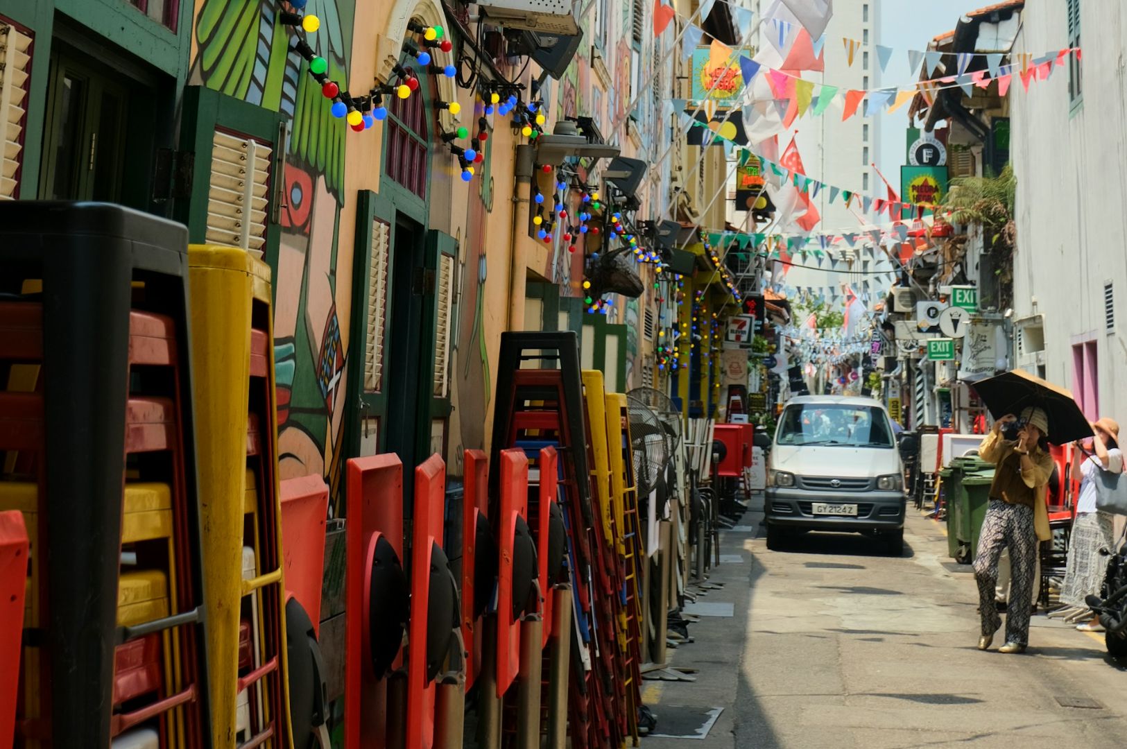 Colorful alley with stacked chairs, painted walls, and hanging lights. Festive flags stretch across, with people and a van adding vibrancy.