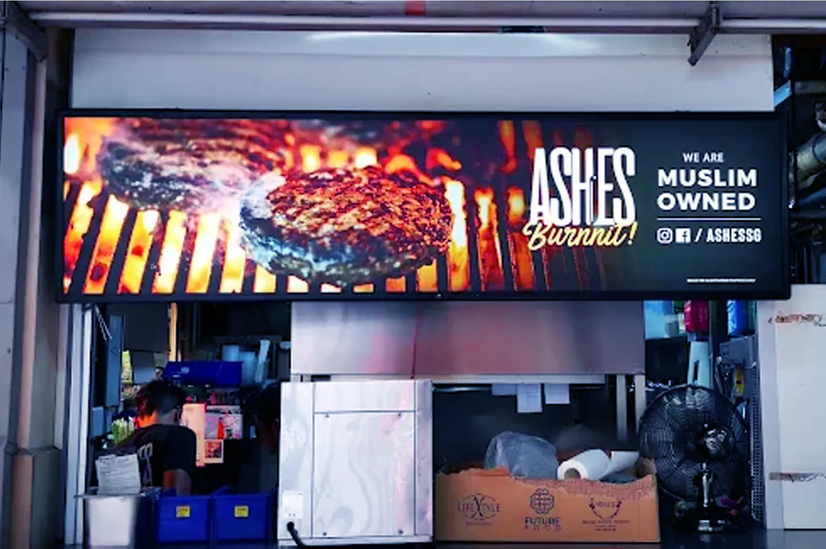 A vibrant sign displays sizzling burger patties on a grill with the words "Ashes Burnnit! We Are Muslim Owned" above a bustling kitchen scene below.