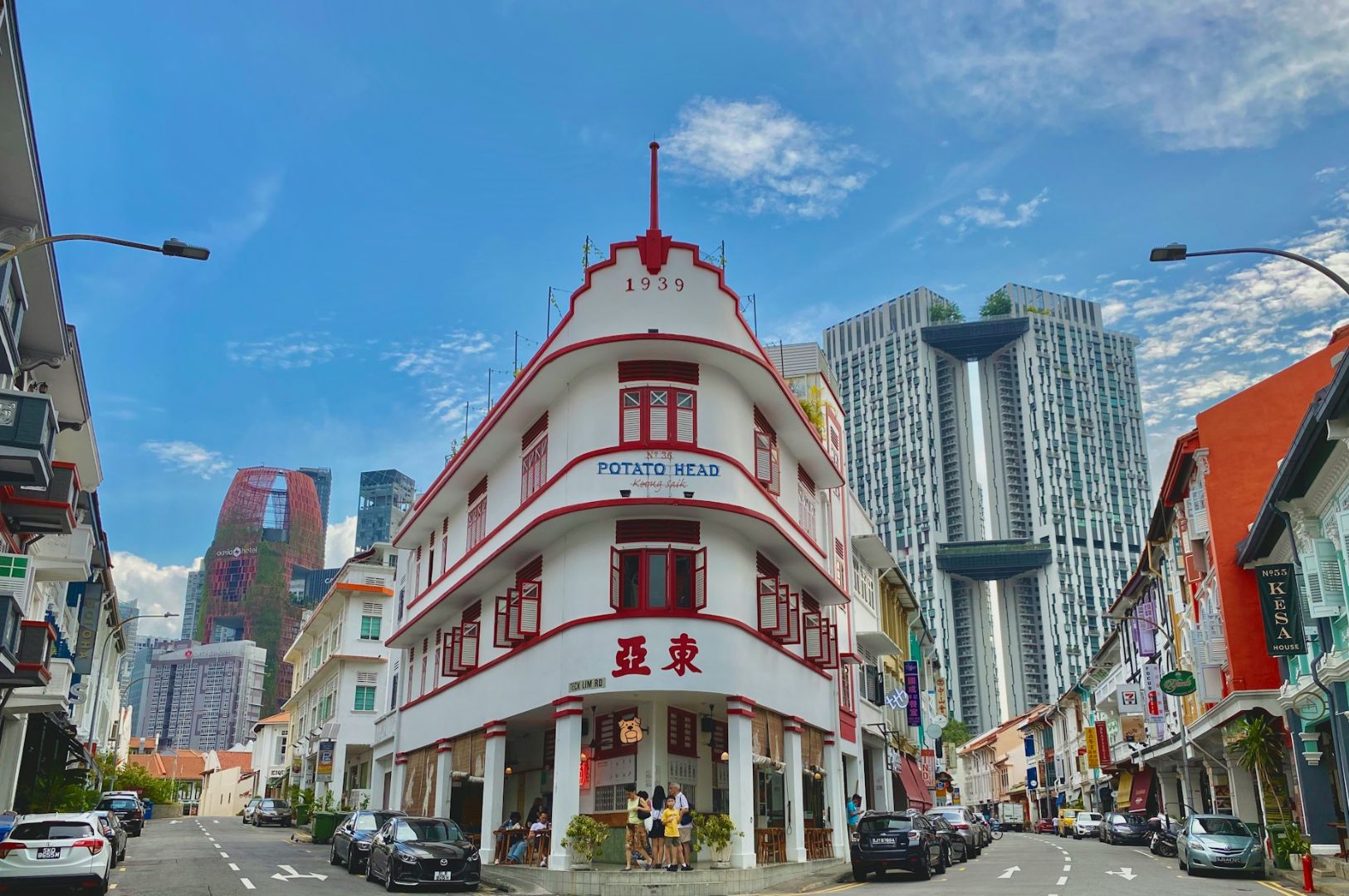 Historic white building with red accents labeled "Potato Head" on a vibrant street. Modern skyscrapers rise in the background under a clear blue sky.