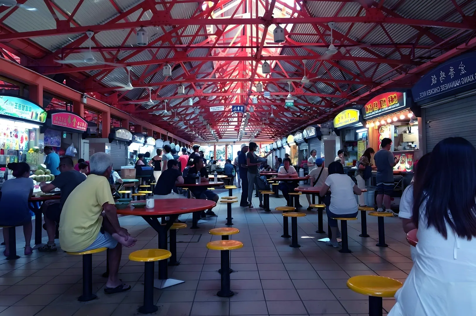 Bustling indoor food market with red metal beams, vendor stalls on both sides, and people seated at tables with vibrant yellow stools under warm lighting.