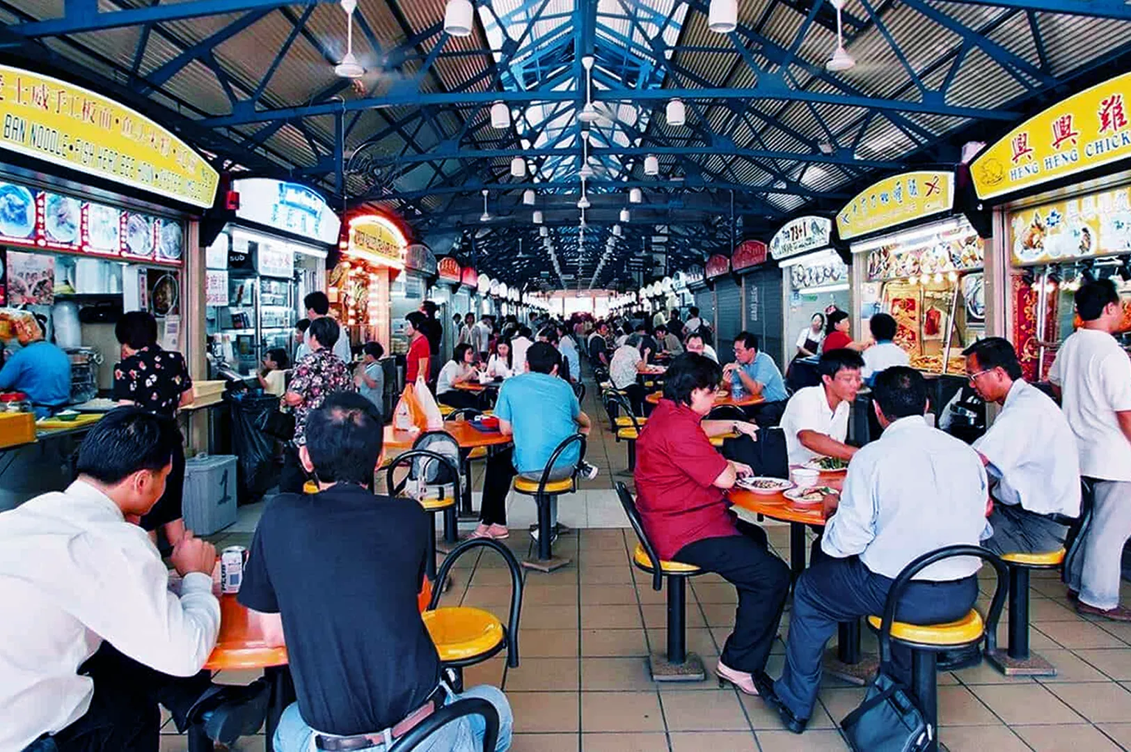 A bustling food court with people sitting at yellow tables under a blue metal roof, surrounded by vibrant food stalls, conveying a lively atmosphere.