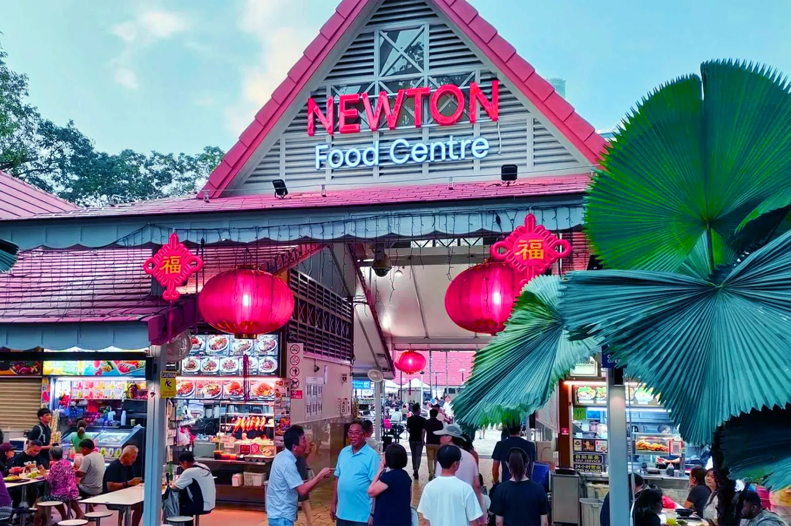 Newton Food Centre bustling with people and stalls under a triangular roof. Red lanterns create a festive vibe. Lush greenery in the foreground.