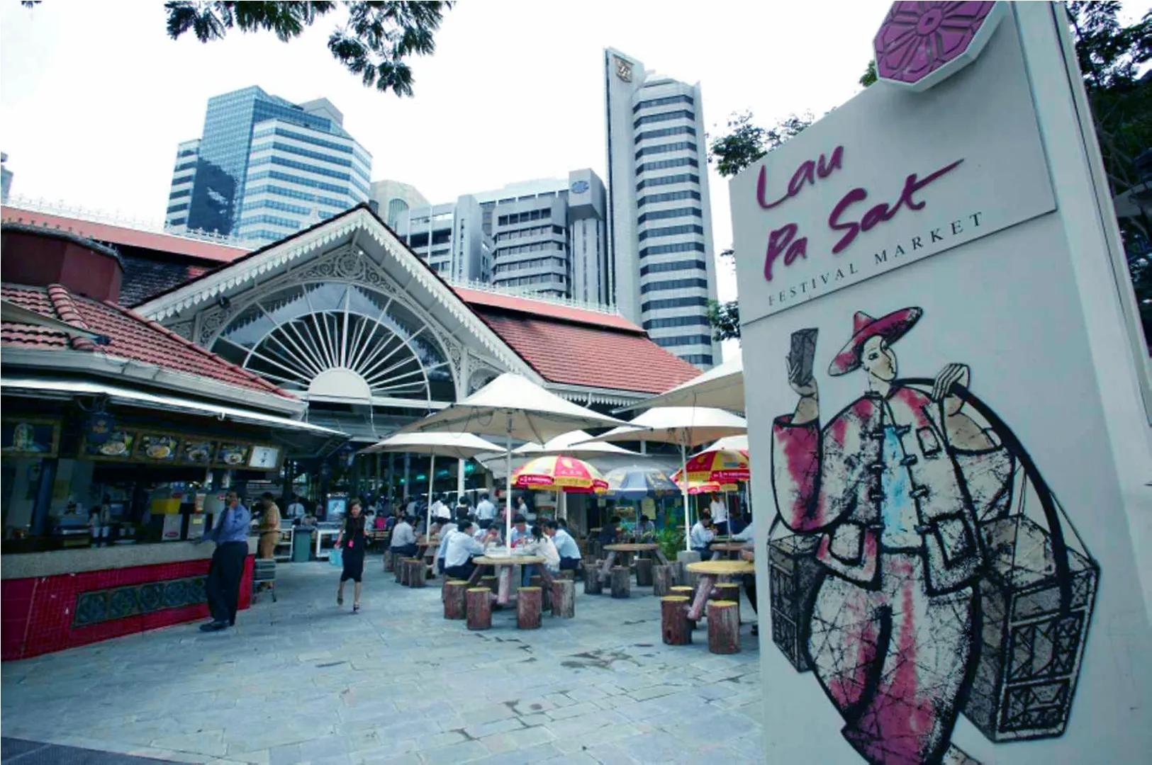 Street view of Lau Pa Sat Festival Market, with people dining under umbrellas. Traditional architecture contrasts with modern skyscrapers in the background.