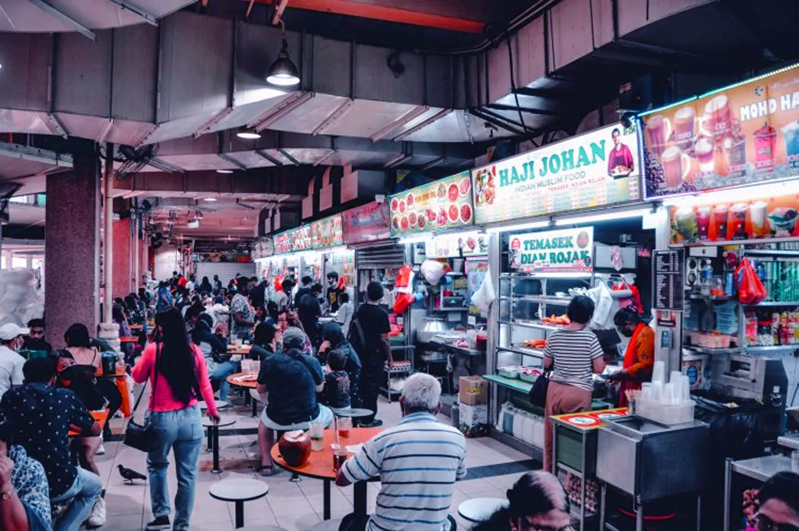 Bustling indoor food court with crowded tables and colorful vendor signs. People of various ages enjoy meals, creating a lively, vibrant atmosphere.