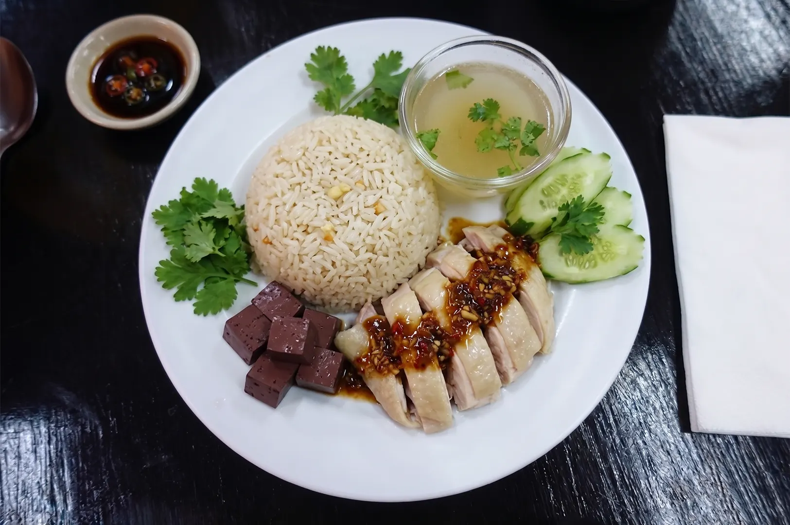 Hainanese chicken rice platter served with sliced poached chicken, seasoned rice, clear broth, cucumber slices, and cubes of chicken blood curd.