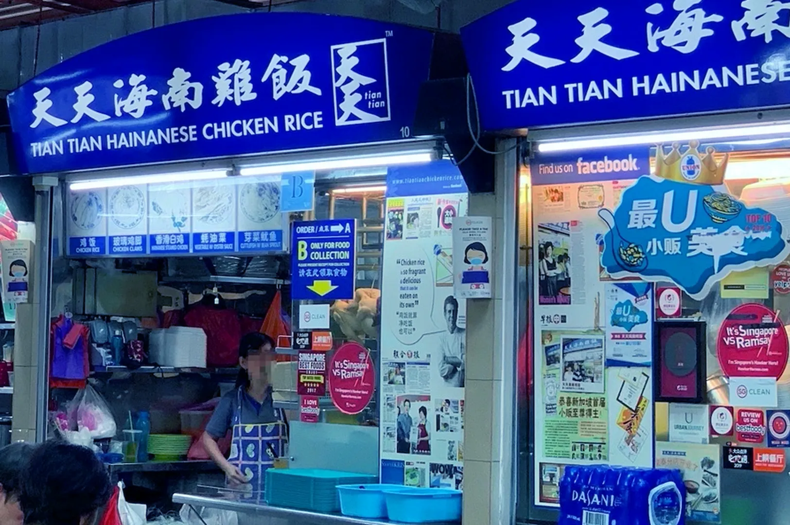Open-air food stall with a blue sign reading "Tian Tian Hainanese Chicken Rice." A staff member is visible inside, surrounded by colorful posters and notices.