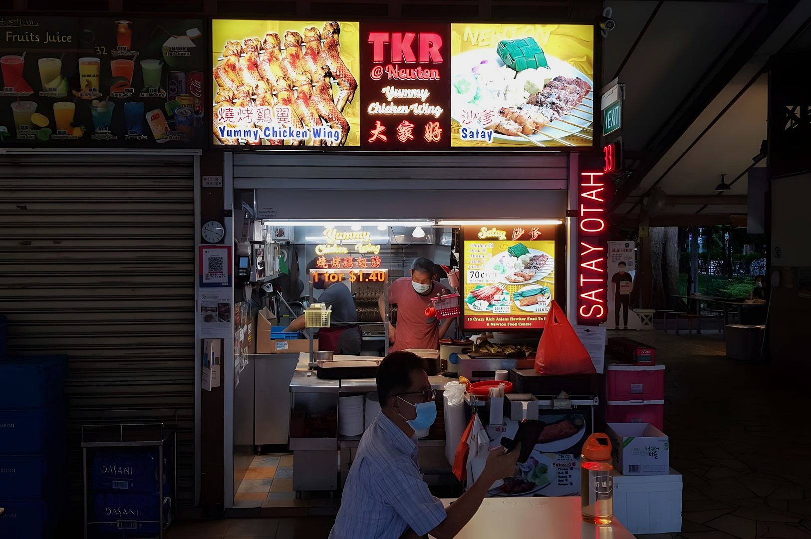 A brightly lit food stall with signs for "Yummy Chicken Wing" and "Satay." A masked vendor prepares food. A customer, also masked, waits in the foreground.