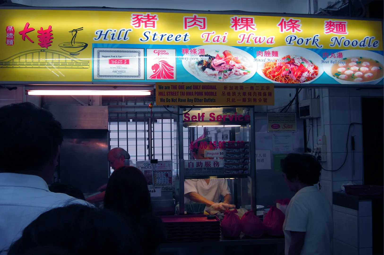 Hawker stall with bright yellow signage reading "Hill Street Tai Hwa Pork Noodle." Displayed images of pork dishes and a self-service counter below.