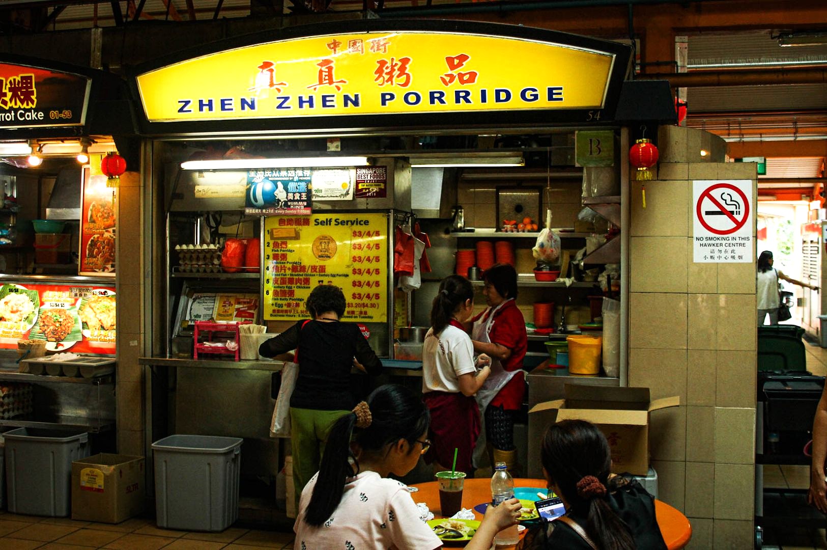 Hawker center stall "Zhen Zhen Porridge" with a yellow sign. Three customers order food under fluorescent lights. A seated woman eats nearby.