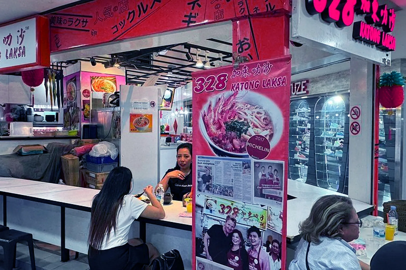 A vibrant indoor food stall with a large "328 Katong Laksa" sign, featuring posters of laksa dishes. Three people are enjoying a meal, creating a lively and cozy atmosphere.