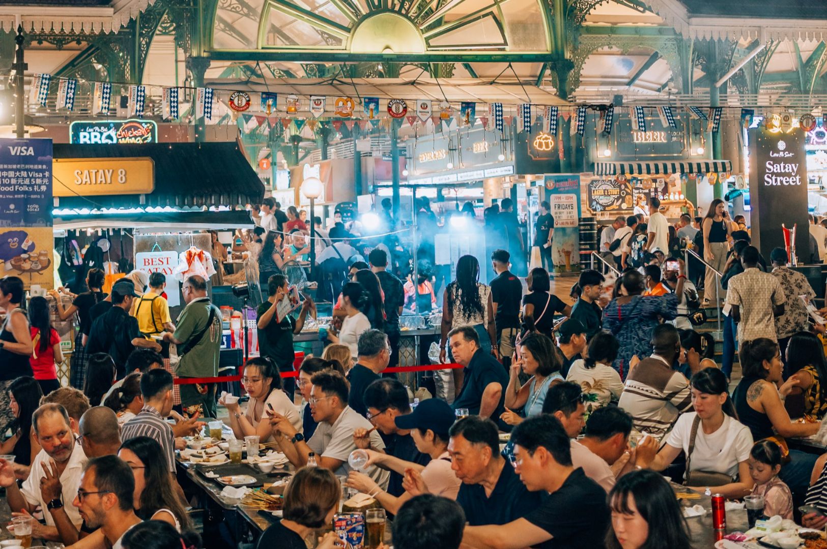 A bustling outdoor food market with crowds enjoying diverse dishes at shared tables. Stalls line the background, colorful and vibrant, creating a lively atmosphere.