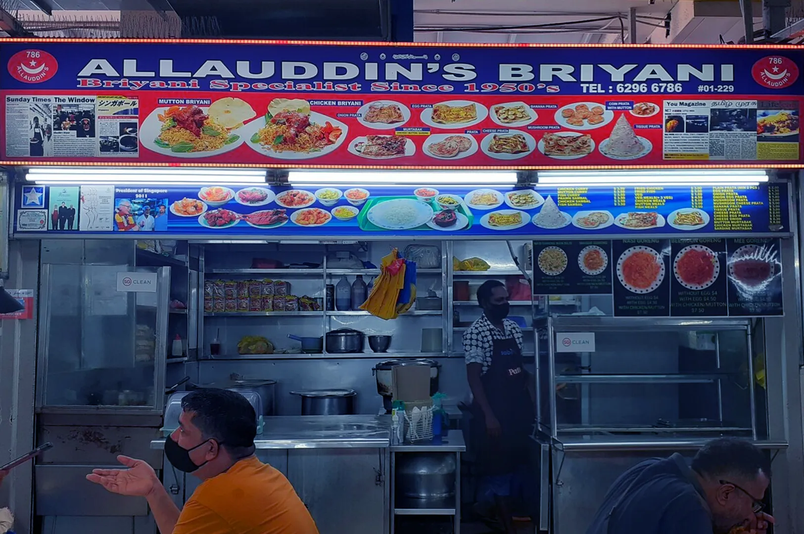 Stall named "Allauddin's Briyani" with a colorful menu display of various dishes. A person wearing an apron stands inside, while a customer in orange sits at the counter.