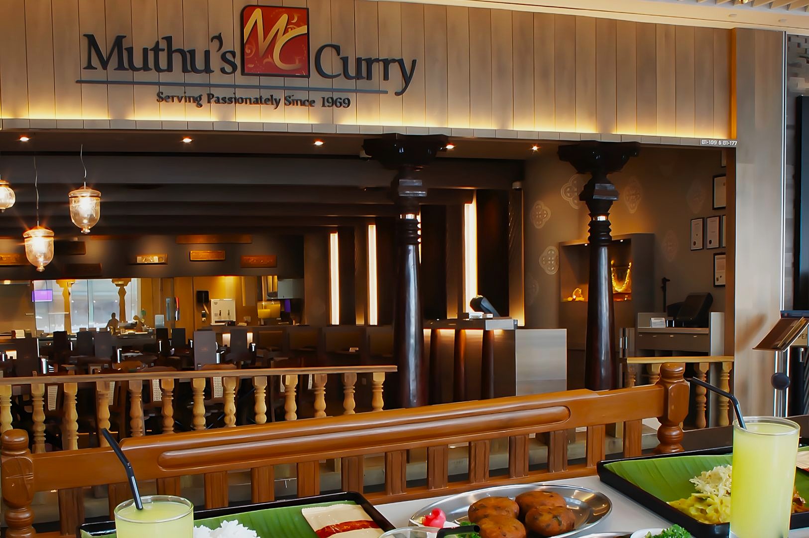 Restaurant interior of Muthu's Curry, stylishly lit, with wooden décor. Indian food and drinks are set on a table, creating a welcoming ambiance.