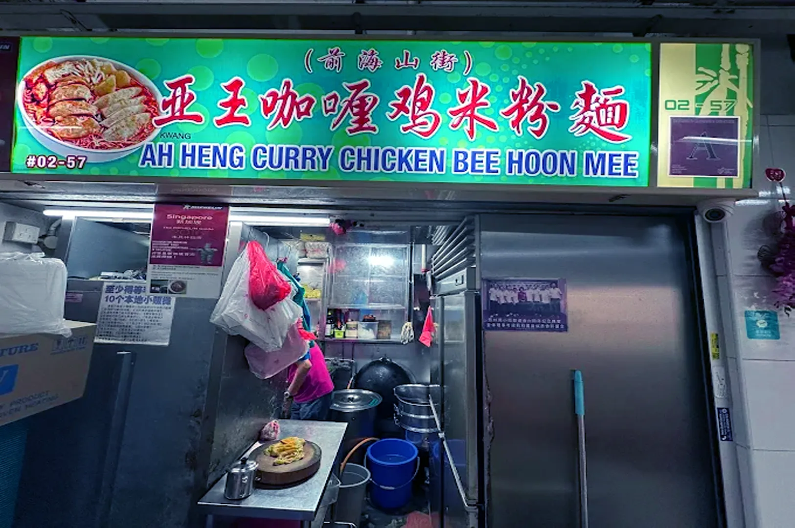 Brightly lit food stall with a green sign reading "Ah Heng Curry Chicken Bee Hoon Mee." The stall appears busy, with cooking utensils and vibrant decor.