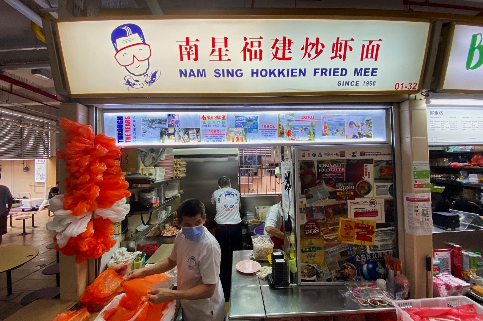 Hawker stall "Nam Sing Hokkien Fried Mee" with bright signage. Two people prepare food, one in a mask. Vibrant scene with a busy, welcoming feel.
