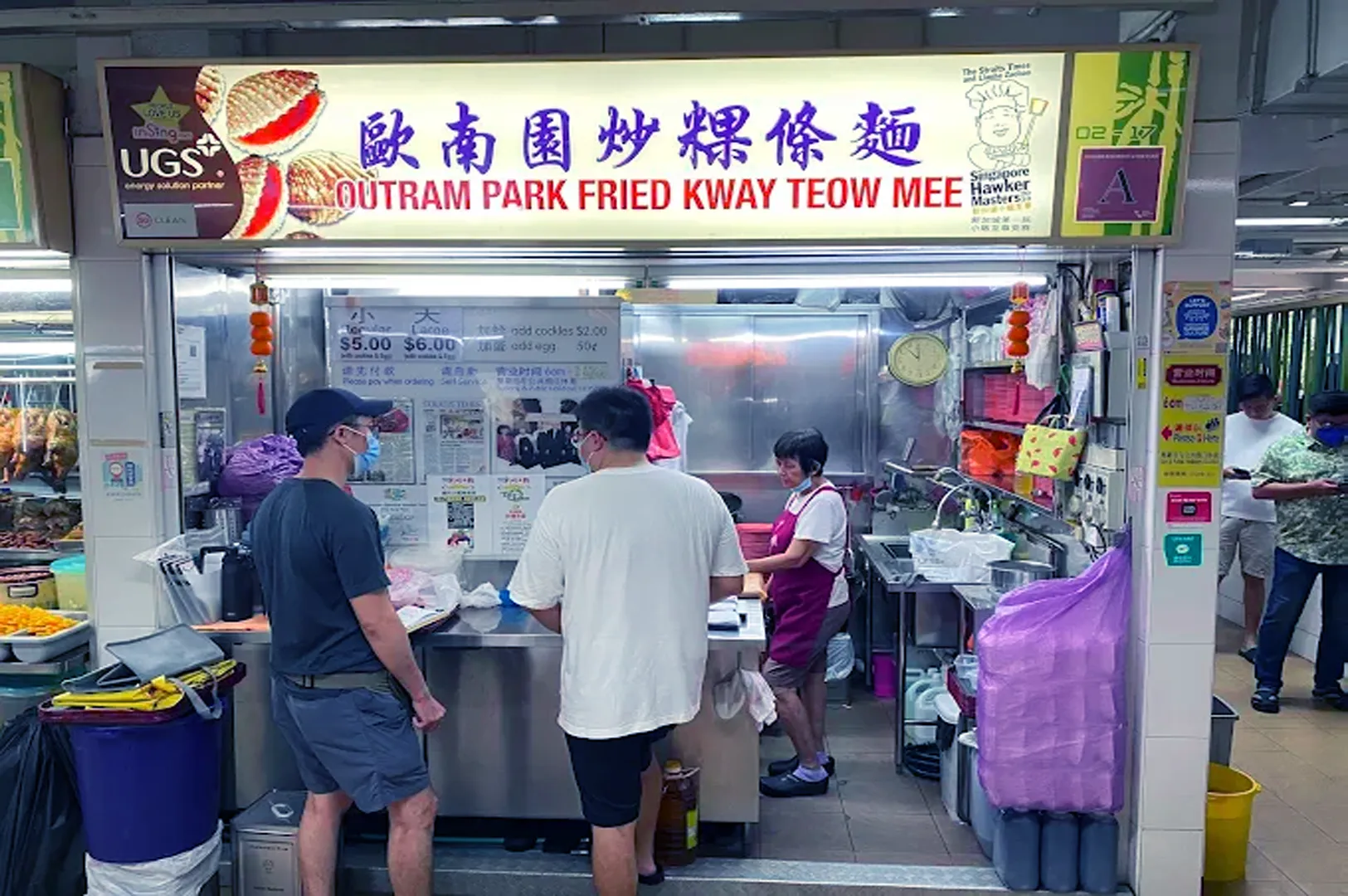 A small, bustling hawker stall named Outram Park Fried Kway Teow Mee. Two customers stand in front, while a vendor in an apron prepares food inside.
