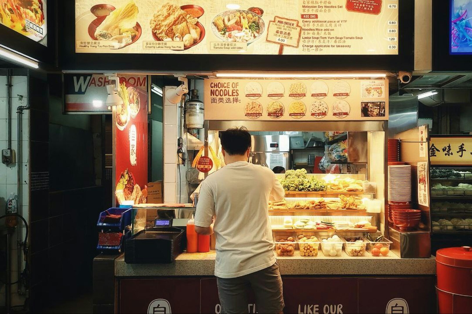 A man stands at a noodle shop counter inside a food court. Bright lights illuminate the display of ingredients. Menus are visible above. Cozy atmosphere.