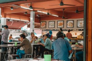 A bustling food court with people seated at tables, enjoying meals. Bright decor, food pictures on the wall, and a lively, social atmosphere.