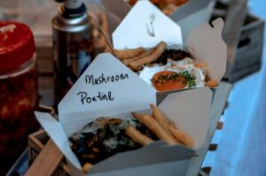 Takeout boxes filled with fries and topped dishes, labeled "Mushroom Poutine." A sunny-side-up egg garnishes one, set against a casual dining backdrop.