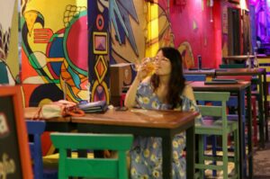 A woman in a patterned dress sips a drink at a rustic table, surrounded by colorful, vibrant murals in a lively, artistic alley. Tables and chairs add to the casual, eclectic atmosphere.