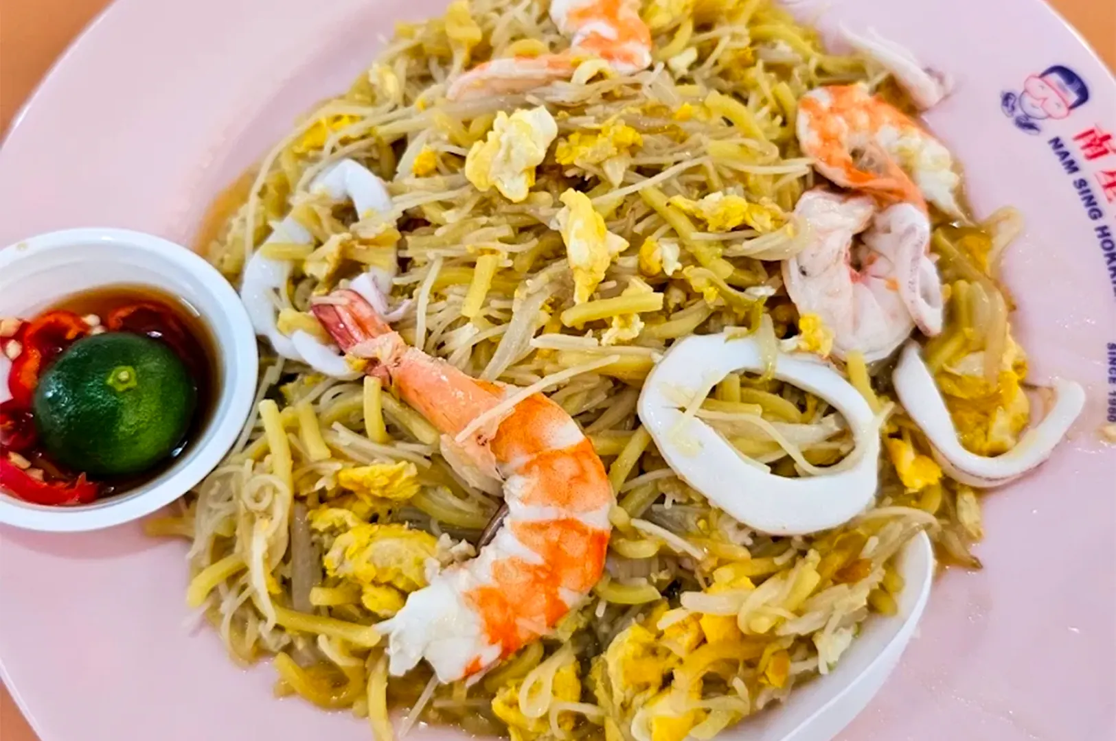 A plate of Nam Sing Hokkien Fried Mee, a classic Singaporean dish featuring stir-fried noodles with shrimp and squid, served with chili and lime on the side.