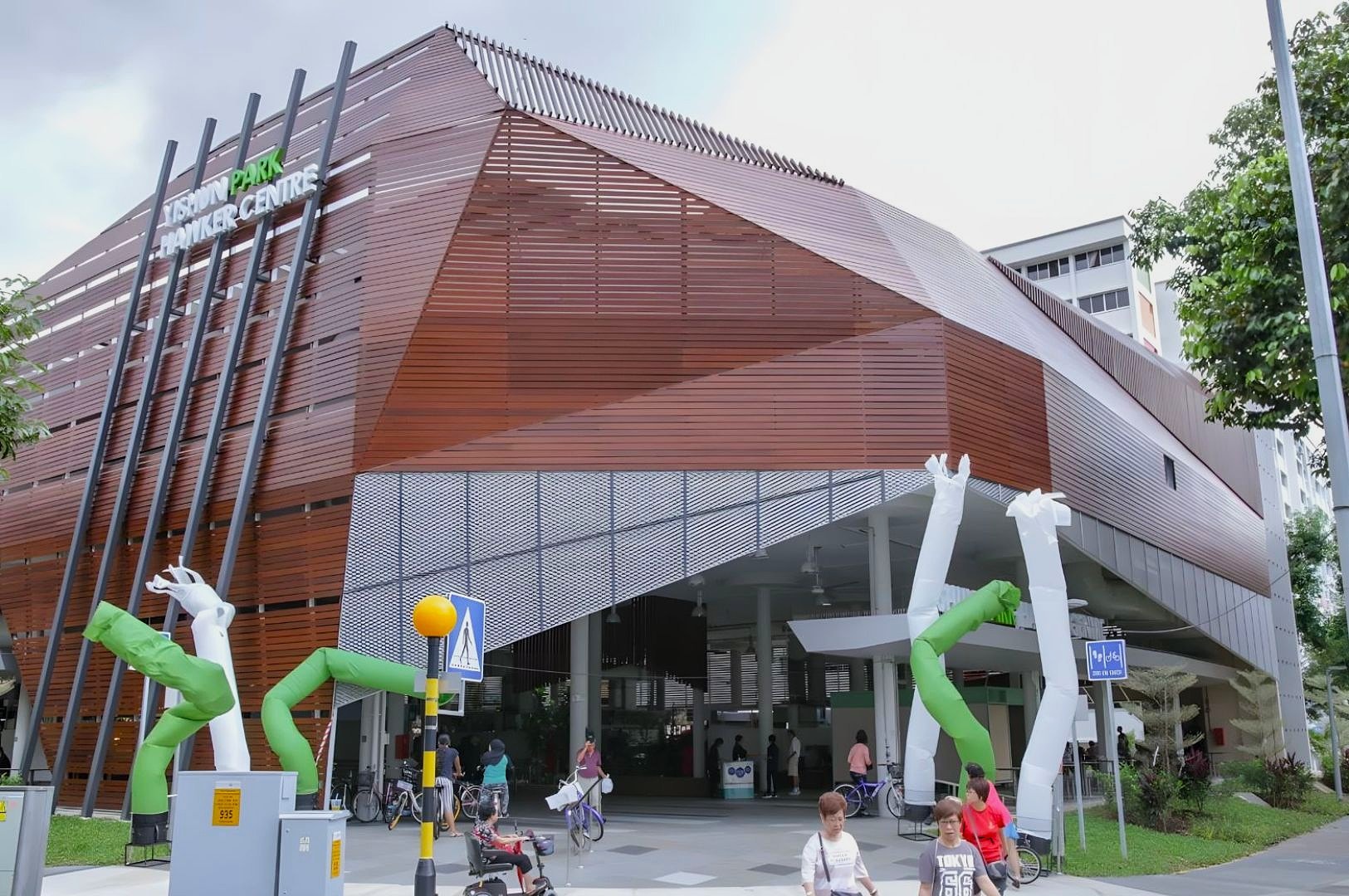 Modern building with a striking angular facade, marked as "Teban Park Hawker Centre." White and green inflatable tube men add a playful touch outside.