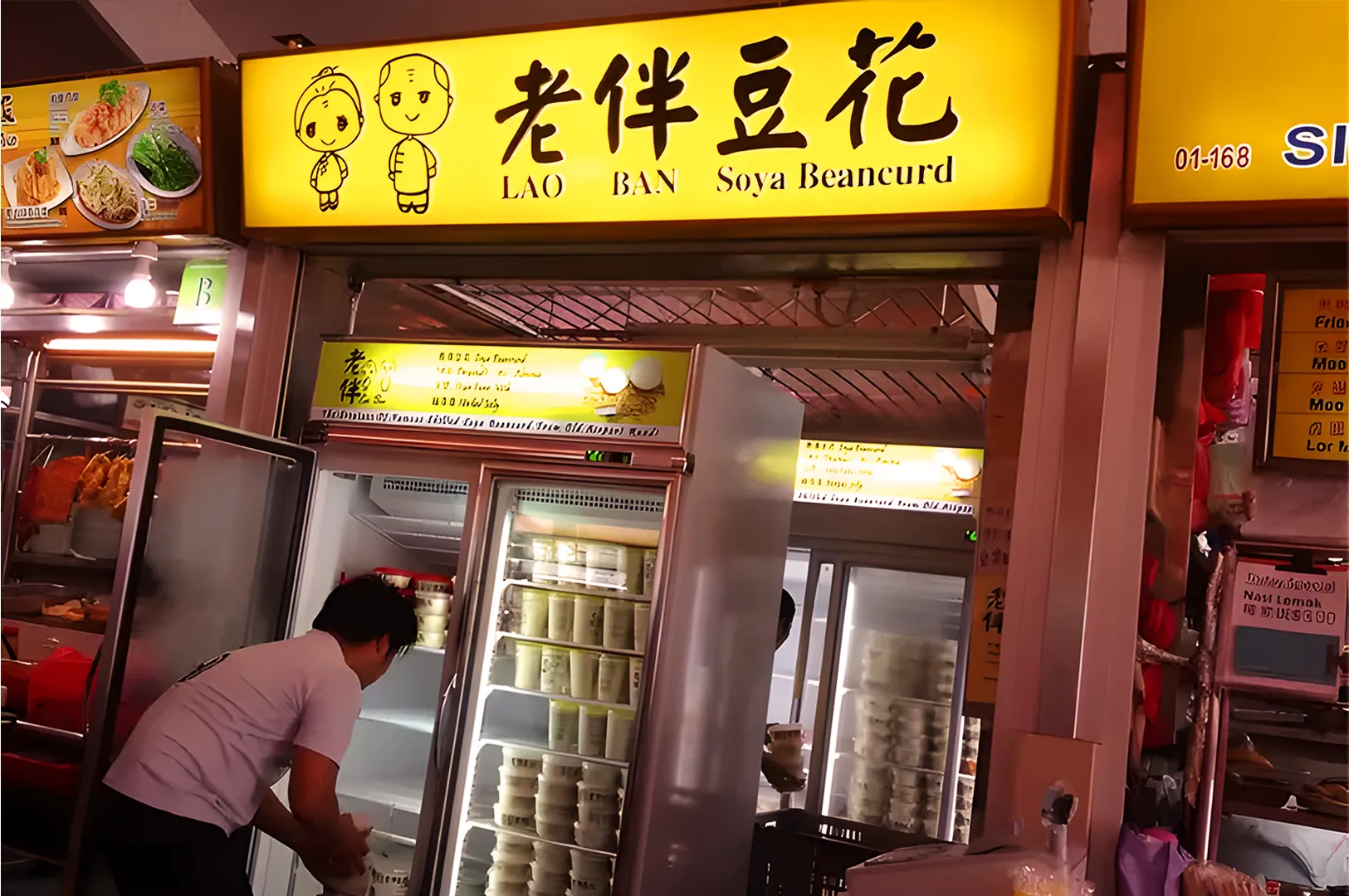 Brightly lit food stall with "Lao Ban Soya Beancurd" sign. Fridge doors open, shelves filled with beancurd containers. Worker stacks items, creates a busy, inviting atmosphere.