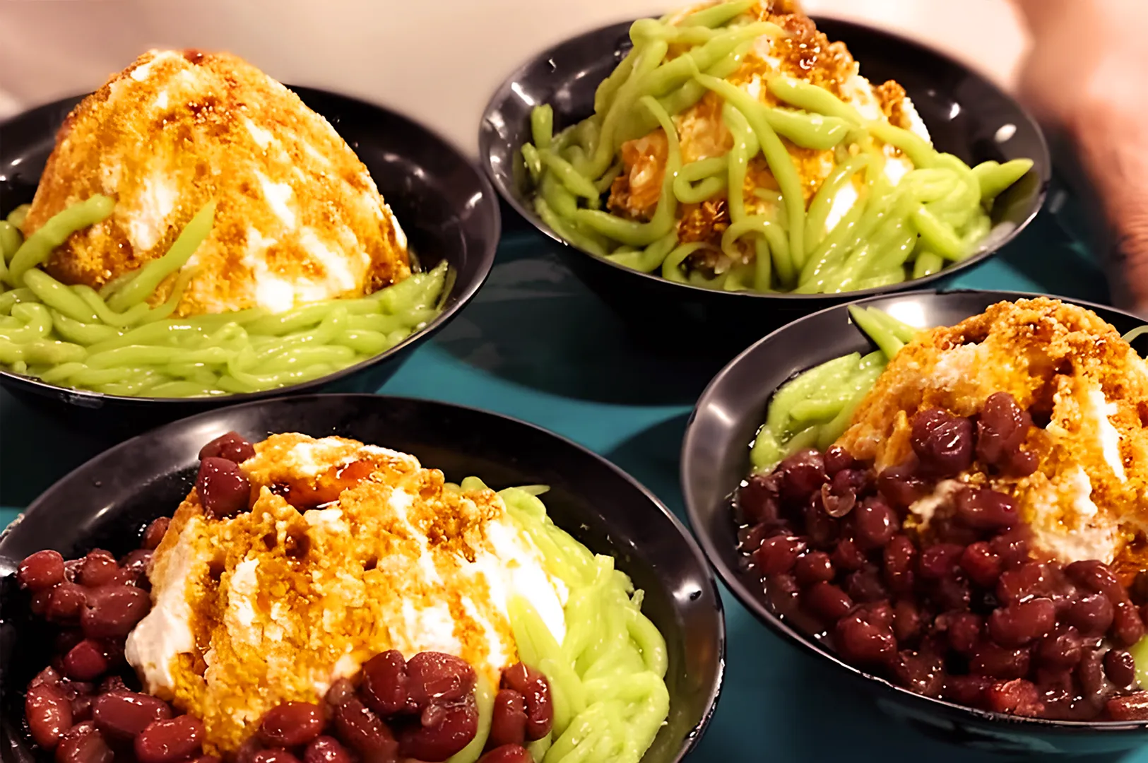 Four bowls of dessert with shaved ice, topped with green pandan noodles, sweet red beans, and drizzled with palm sugar syrup, set on a teal tray.