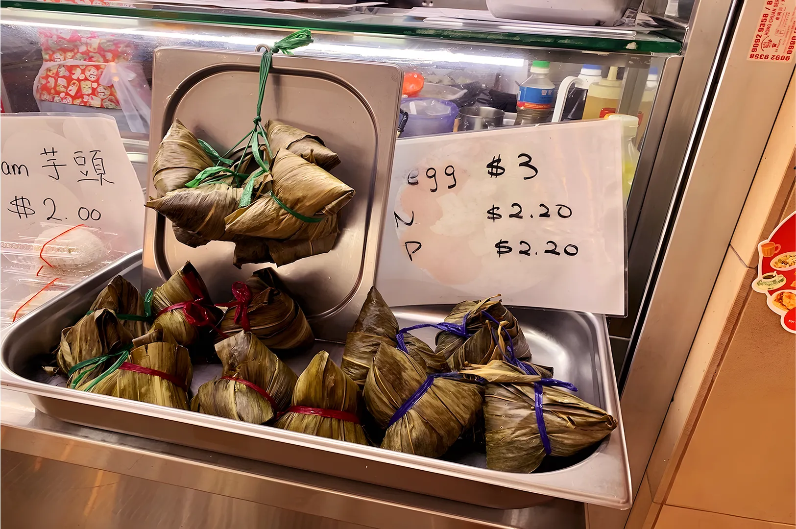 Bundles of rice wrapped in green leaves tied with colored strings are displayed in a metal tray. Price signs show $2.00 and $3.00 options.