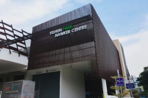 Front of Yishun Park Hawker Centre with wooden slat facade under a blue sky. Nearby, a truck and road signs are partially visible.