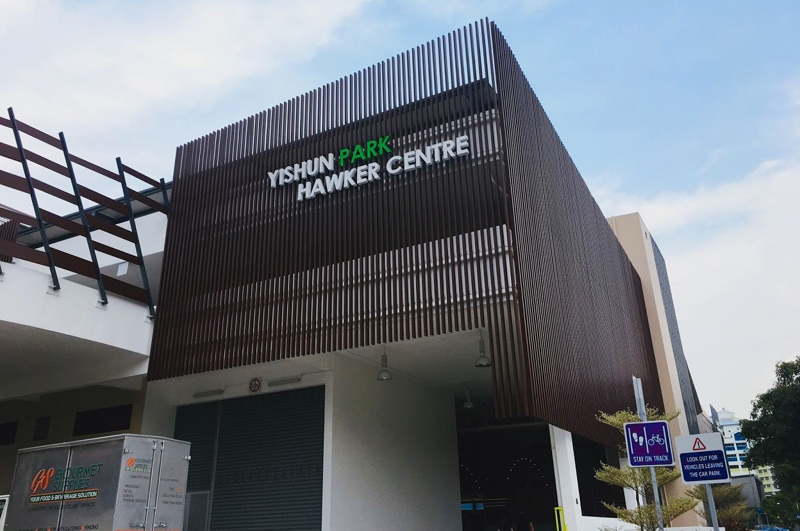 Front of Yishun Park Hawker Centre with wooden slat facade under a blue sky. Nearby, a truck and road signs are partially visible.