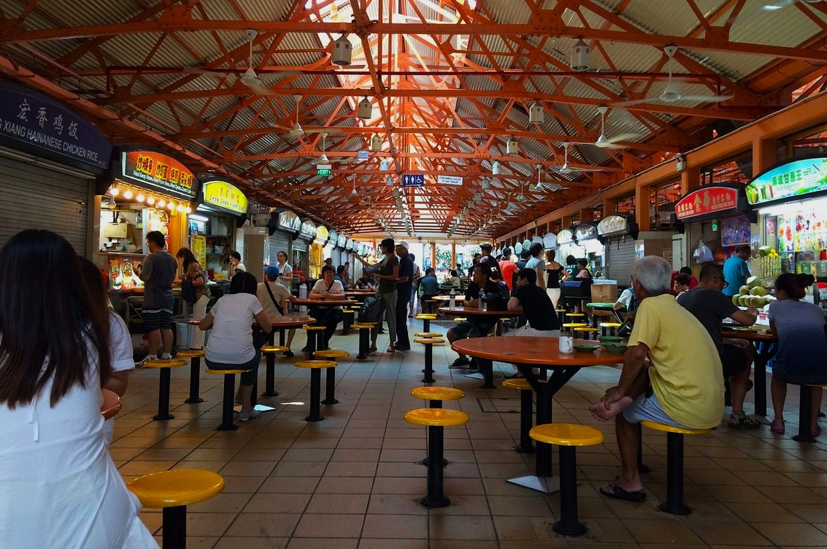Bustling hawker center with people seated at shared tables under a high, red metal roof. Surrounding stalls display illuminated signs and food items.