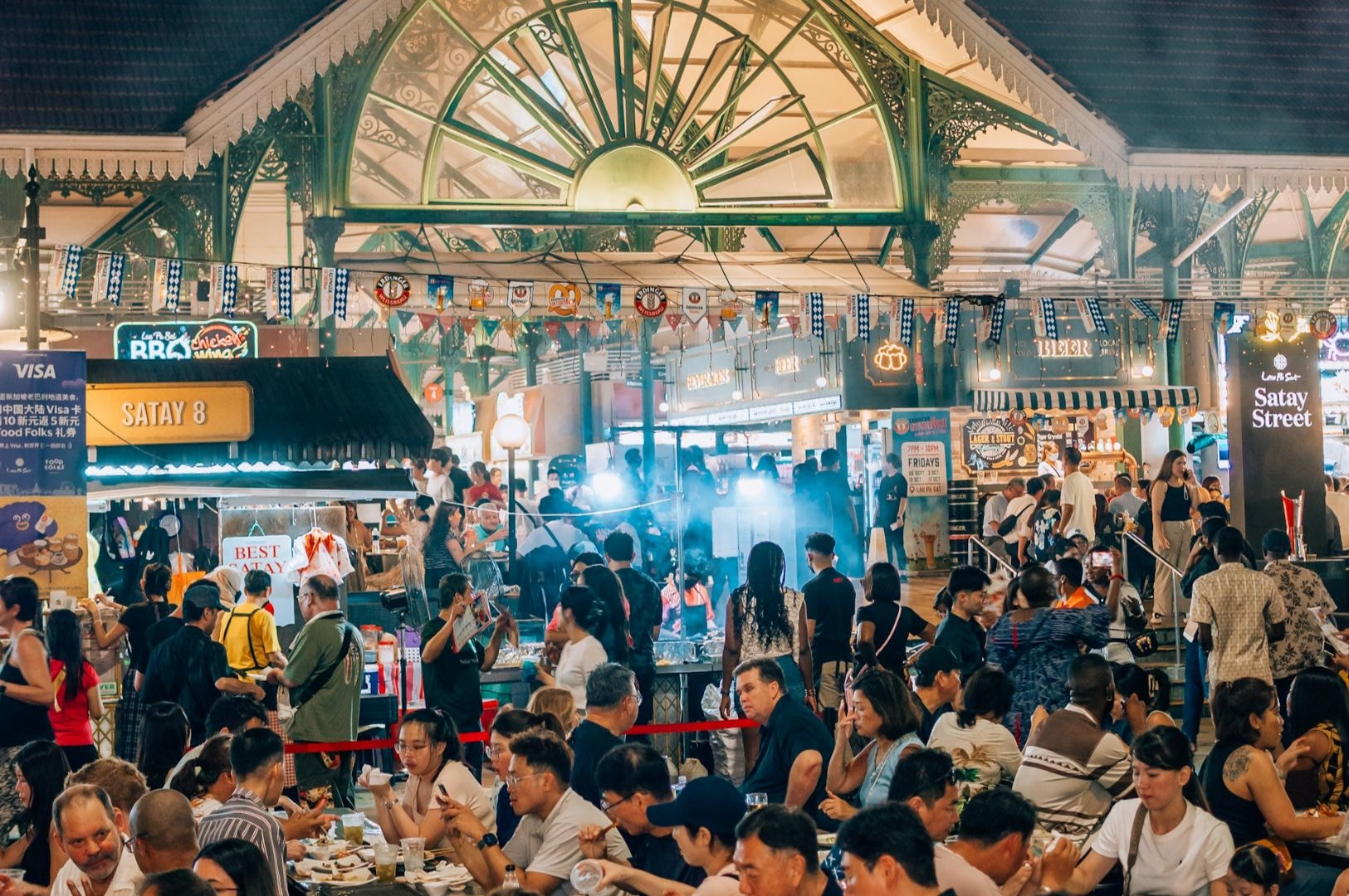 Bustling night market scene with people enjoying food at crowded tables under ornate green arches. Bright stalls, festive flags, lively and vibrant atmosphere.