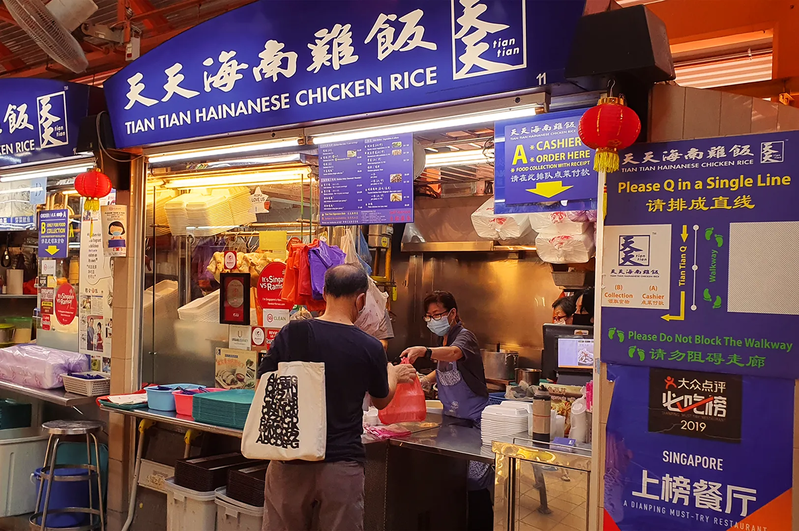 Eye-level wide-angle street photo of Tian Tian Hainanese Chicken Rice hawker stall, showing customers ordering at the counter under bright blue signage and menu boards inside a busy Singapore food market.