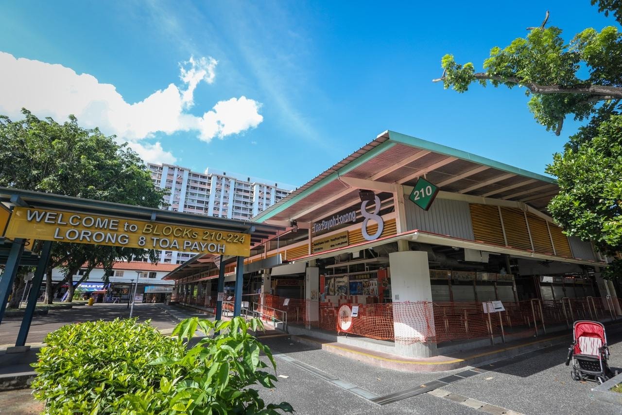 This image shows the exterior of the Toa Payoh Lorong 8 Market & Food Centre, which is partially cordoned off by orange safety netting under a bright blue sky. A yellow overhead sign welcomes visitors to Blocks 210-224, while high-rise residential buildings and lush green trees are visible in the surrounding area.