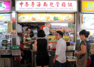  This image captures the busy storefront of Tiong Bahru Hainanese Boneless Chicken Rice, a popular hawker stall featuring a bright menu board with various chicken and vegetable dishes. Customers are seen interacting with the vendor and waiting in line, while whole roasted chickens hang behind the glass display in a typical Singaporean food center setting.