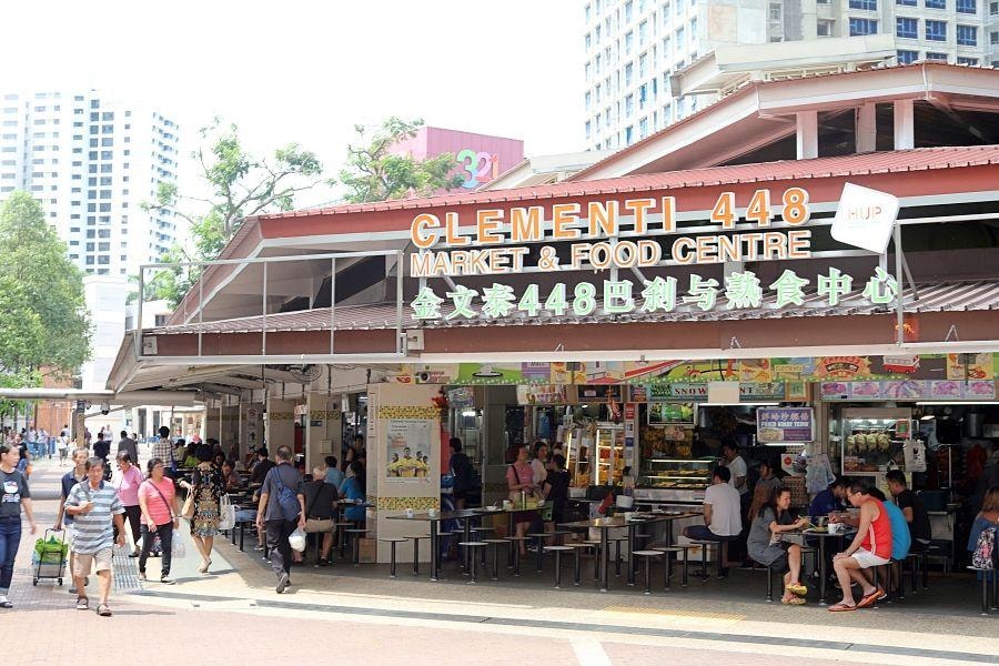  The image shows the bustling entrance of Clementi 448 Market & Food Centre, where patrons are dining at outdoor tables beneath a low-slung, red-tiled roof. The background features modern high-rise residential buildings and a glimpse of the 321 Clementi mall under a bright, clear sky.