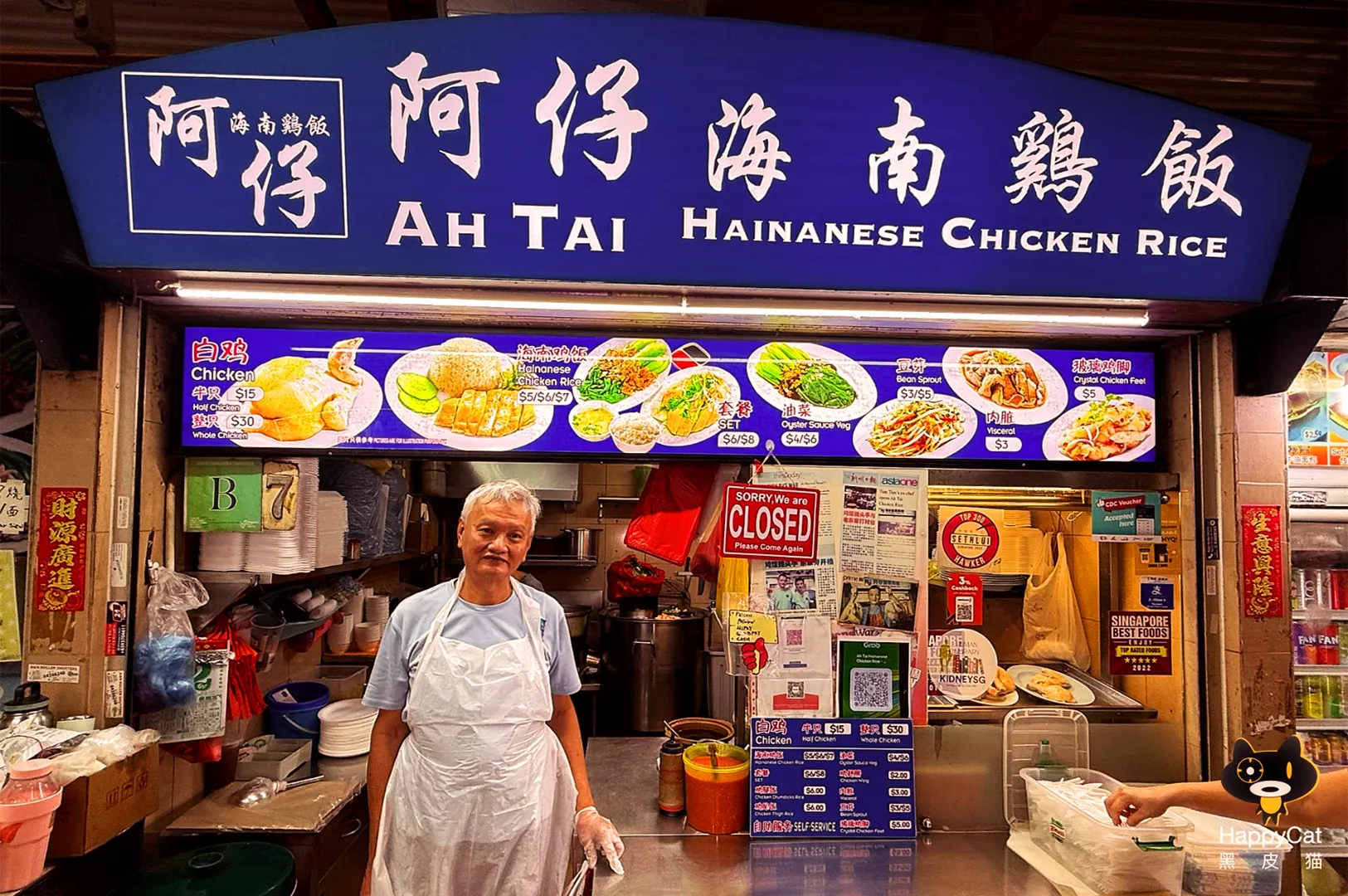 Eye-level frontal wide-angle photo of the Ah Tai Hainanese Chicken Rice hawker stall, featuring an elderly vendor standing behind the counter beneath a blue shop sign with menu photos, captured in a traditional Asian food market setting.